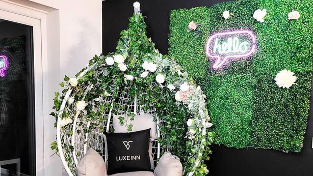 Ein Zimmer mit Hängestuhl, leuchtendem 'hello'-Schild und grüner Wand mit weißen Blumen.
