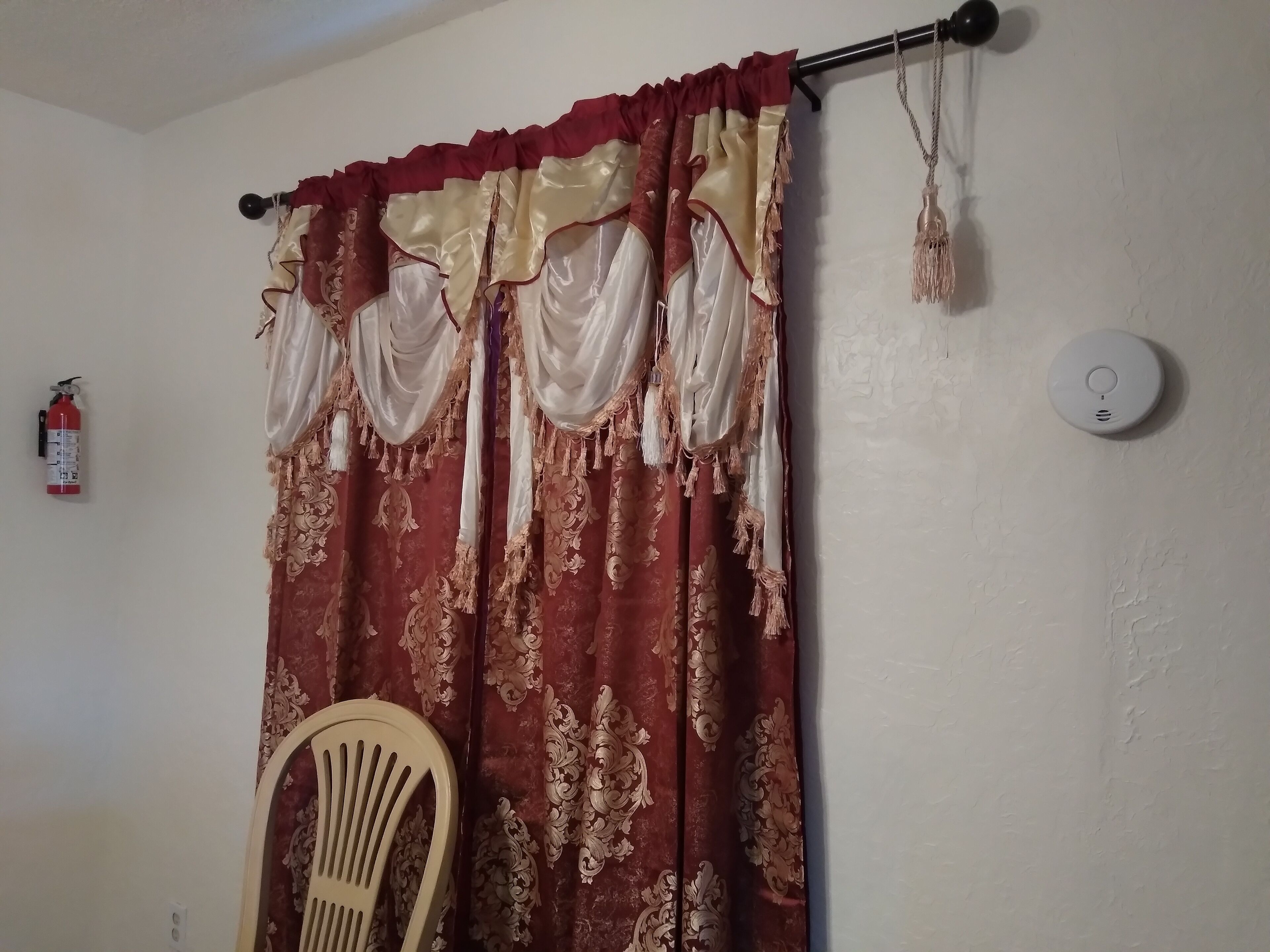 A room with a curtain featuring a floral pattern, a white wall, a fire extinguisher, and a ceiling-mounted smoke detector.