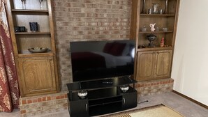 Ein Wohnzimmer mit einem Flachbildfernseher auf einem schwarzen Standfuß, Holzregalen mit dekorativen Gegenständen, einem Kamin aus Ziegelsteinen und einem gemusterten Teppich.