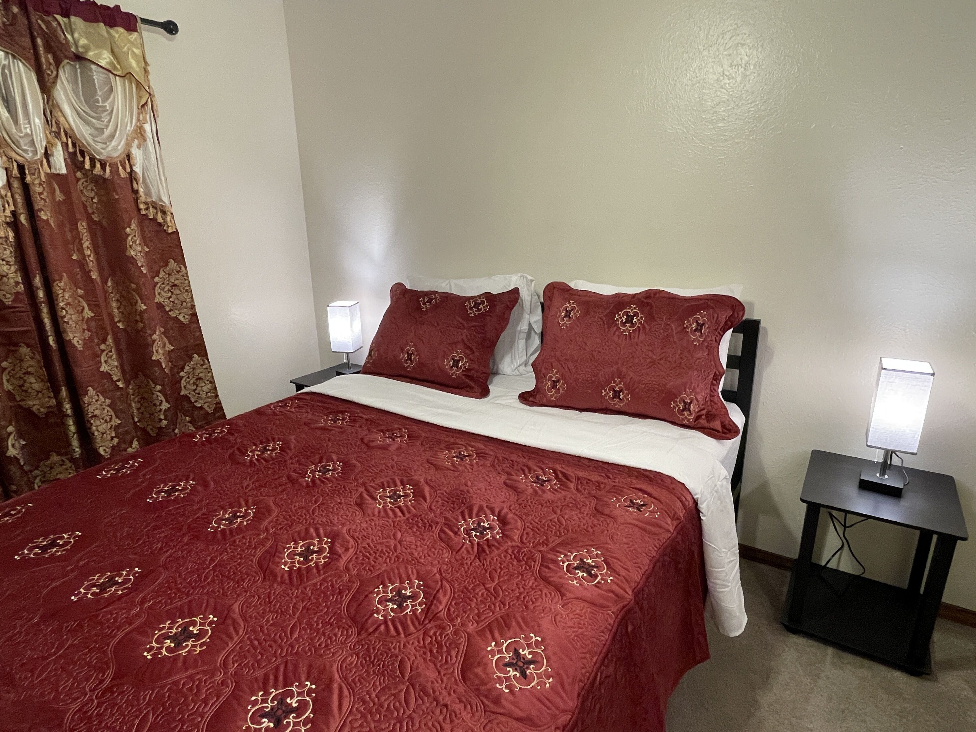 A neatly made bed with a red bedspread and matching pillows, two bedside tables with lamps, and a curtain with a gold pattern.