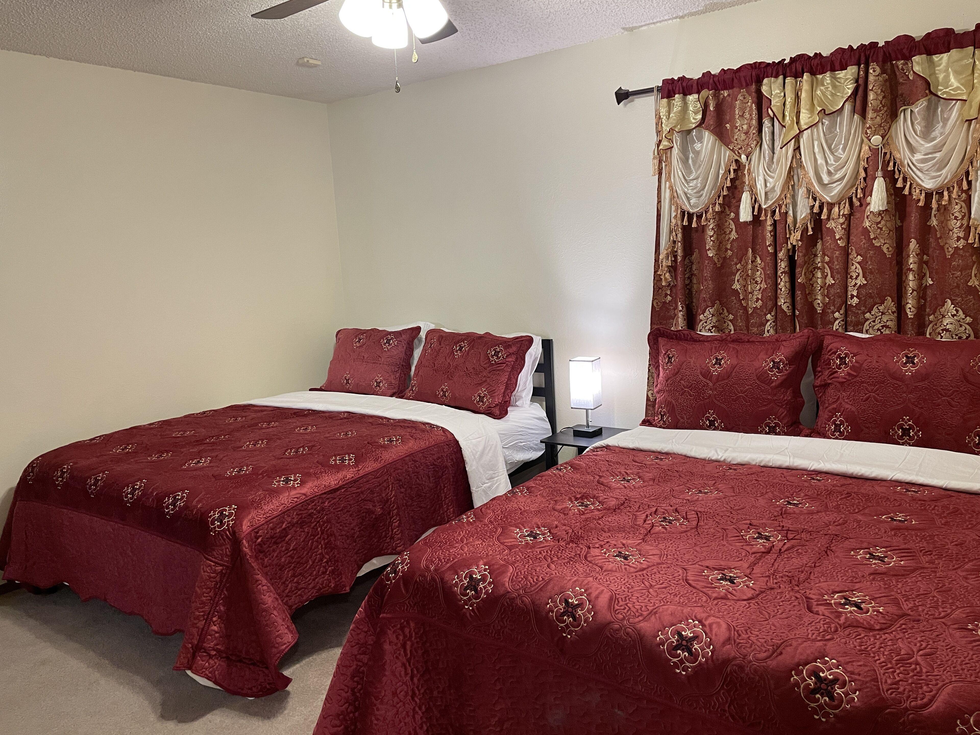 A hotel room with two beds, each with red bedding and white sheets, a ceiling fan, and a window with ornate curtains.
