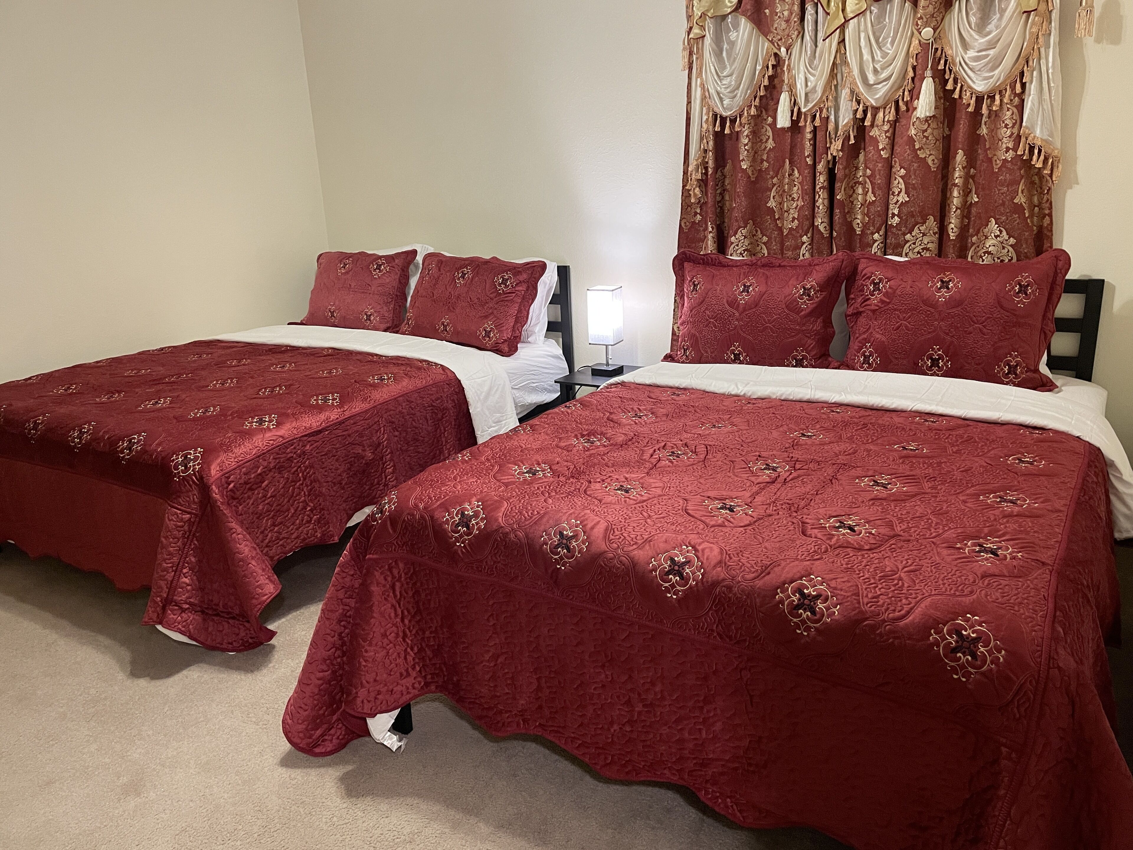 Un lit double avec une couvre-lit rouge et des coussins assortis, une table de chevet avec une lampe, et une fenêtre ornée de rideaux décoratifs.