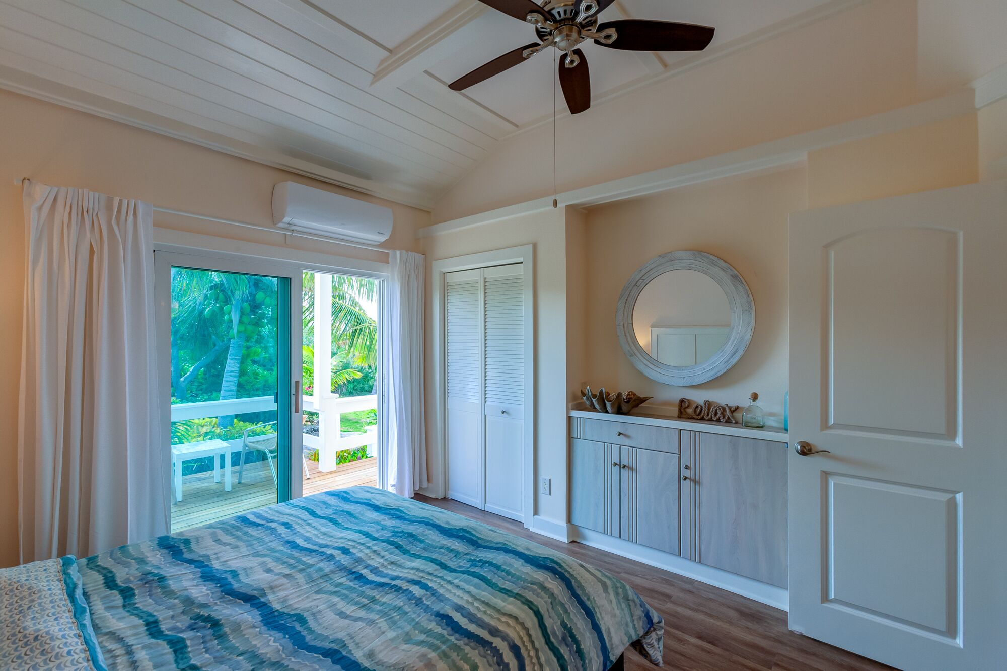 A bedroom with a bed, a ceiling fan, a window with curtains, a door, and a small round mirror on the wall.
