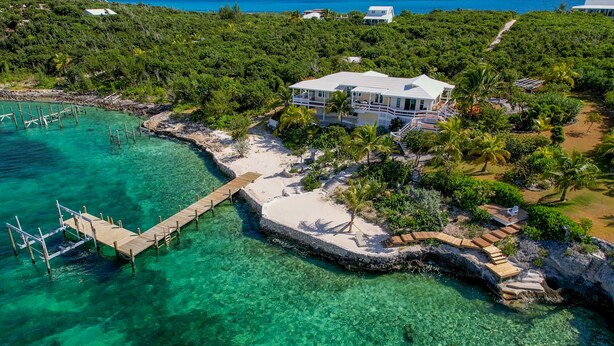 Ein Küsteneigentum mit einer Holzsteganlage, die in klares türkisblaues Wasser führt, umgeben von üppiger Vegetation und einem weißen Haus mit einem Balkon.