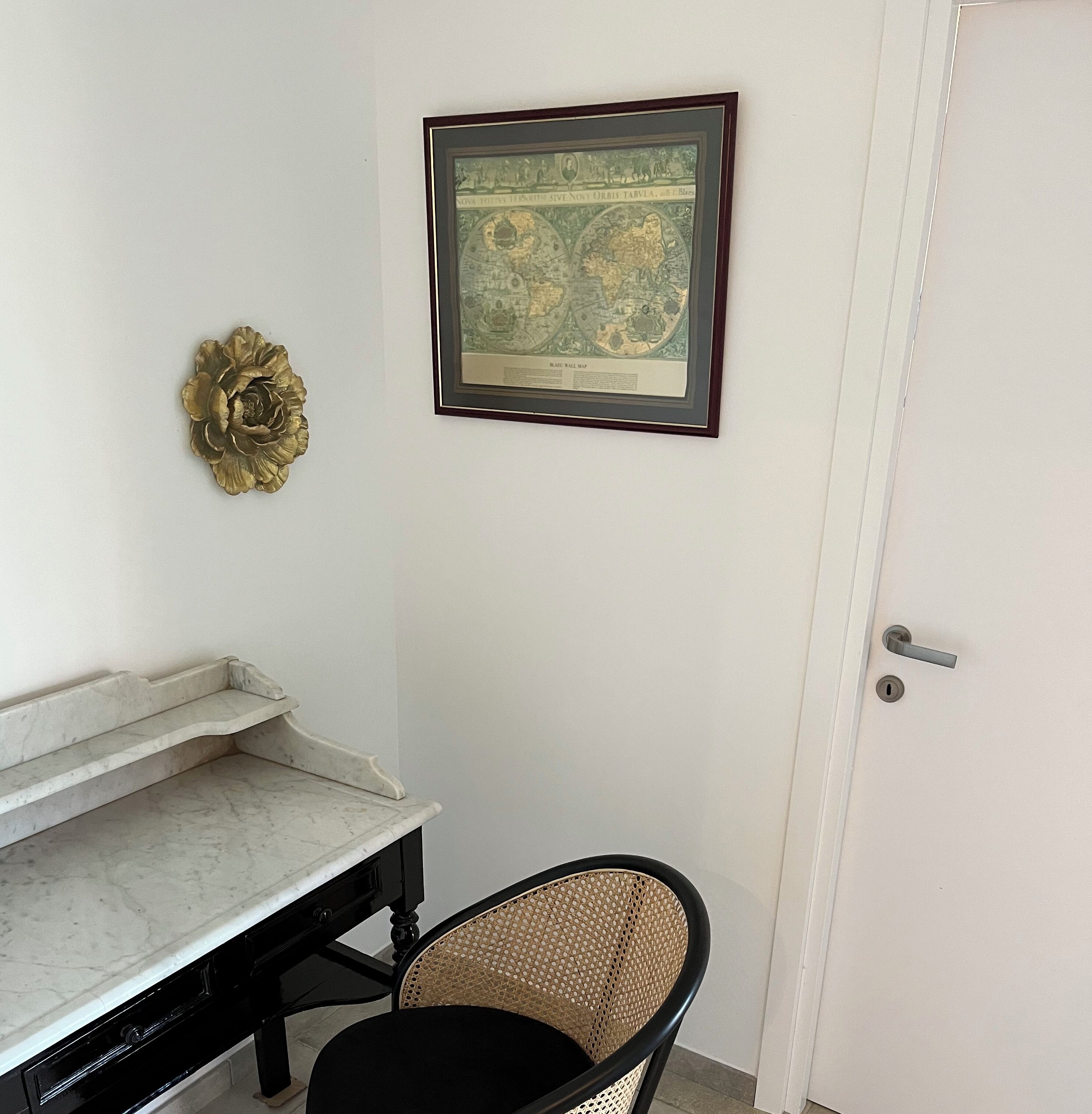 Una habitación con una puerta blanca, un mapa enmarcado en la pared, una mesita con encimera de mármol y una silla de mimbre.