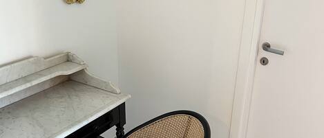 Una habitación con una puerta blanca, un mapa enmarcado en la pared, una mesita con encimera de mármol y una silla de mimbre.
