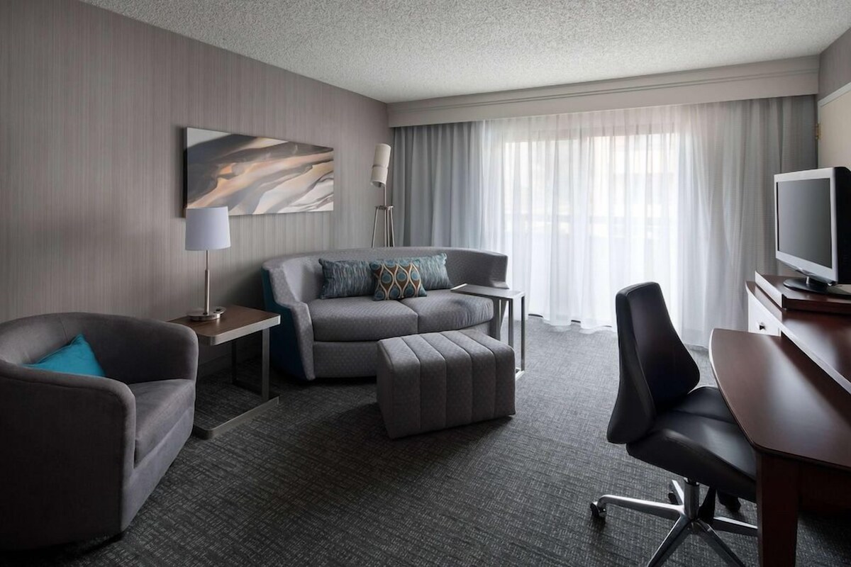A modern hotel room with a grey sofa, a matching ottoman, a desk with a television, and a floor lamp.