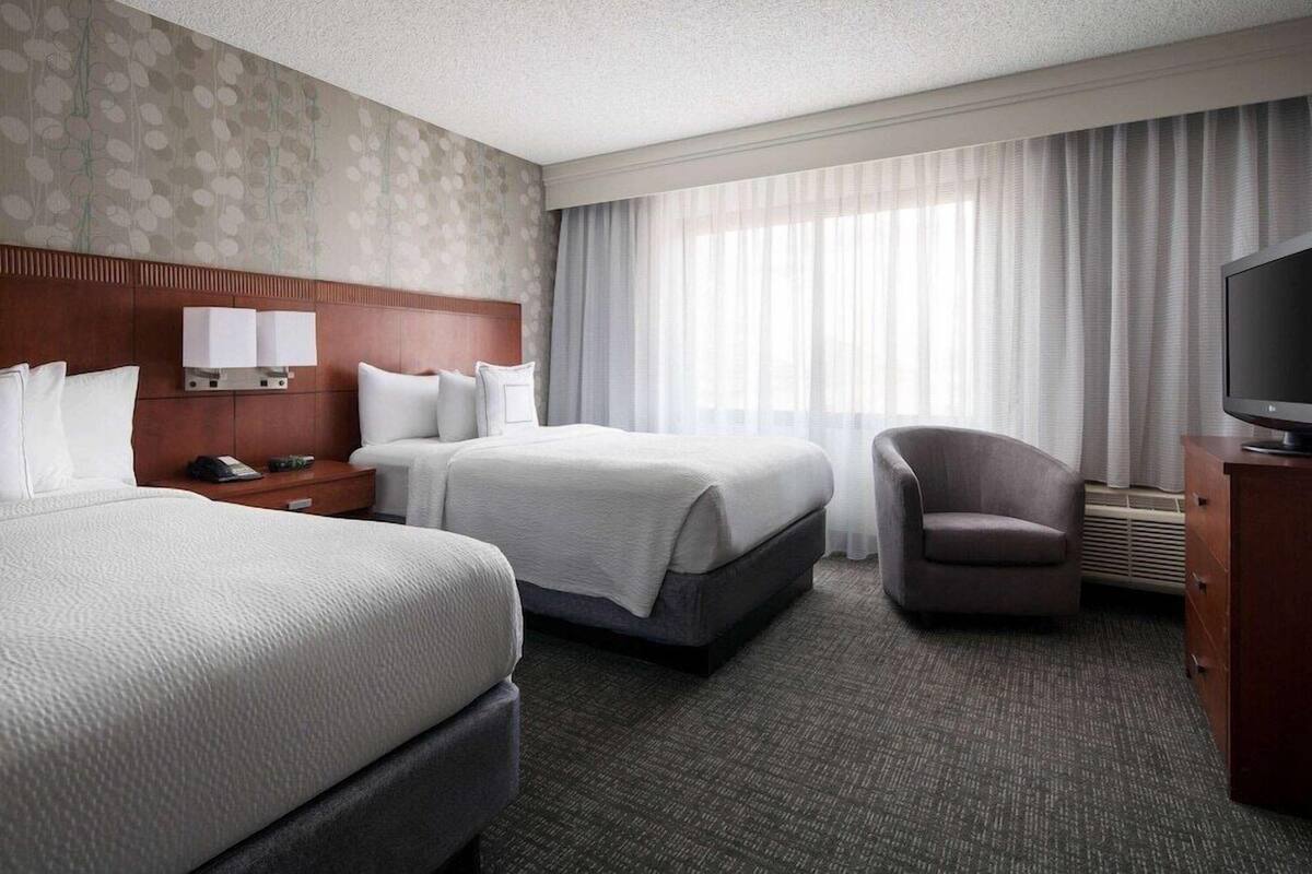 A hotel room with two beds, a TV, a chair, and a desk with a lamp.