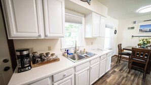 A kitchen with white cabinets, a stainless steel sink, and a coffee maker. There is a window with blinds, a wooden dining table with chairs, and a clock on the wall. - 2 E-Bikes & Kayaks included & No Cleaning Fees!! (Clear Lake)