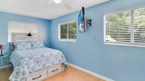 Une chambre à coucher comprenant un lit, une table de chevet, un téléviseur fixé au mur et deux fenêtres équipées de stores.