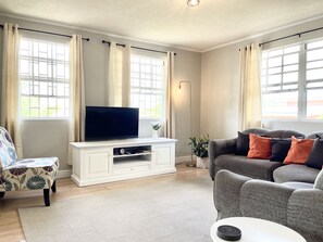 Un salon avec un téléviseur à écran plat posé sur un meuble de télévision blanc, un canapé gris agrémenté de coussins orange et noirs, un fauteuil à motifs et une table basse blanche.