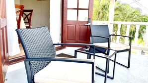 A balcony with two wicker chairs and a table, a wooden door, and a view of greenery.