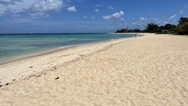 A wide sandy beach with clear turquoise water, a lighthouse in the distance, and a few people walking.