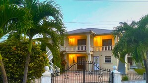 A two-story house with a balcony, a gated entrance, and a well-maintained garden with palm trees.