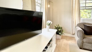 A modern living room with a flat-screen TV on a white console, a grey sofa, a floor lamp, and a potted plant.