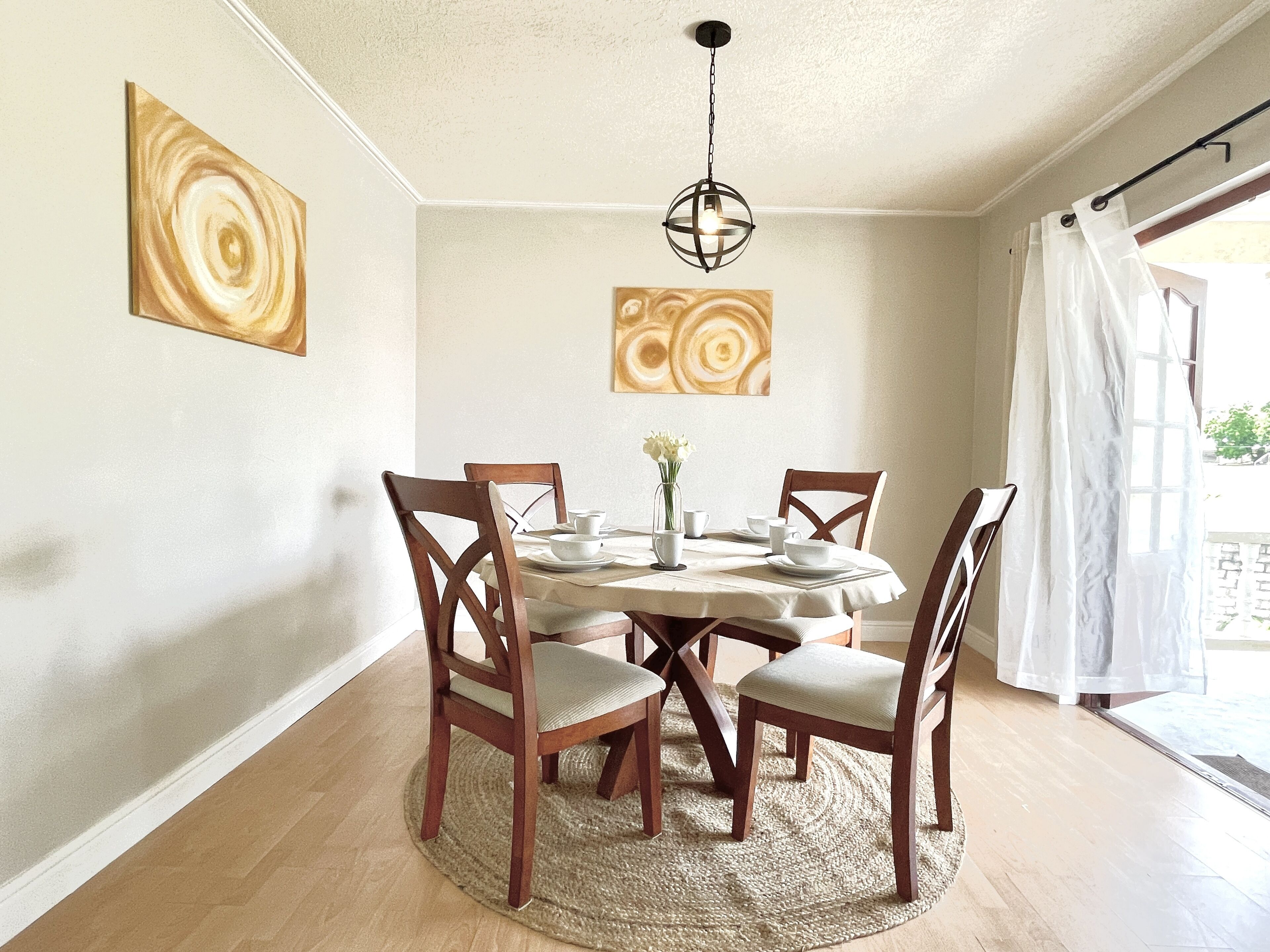 A dining area with a round table, four wooden chairs, a woven rug, and two abstract paintings on the walls.