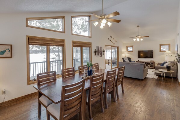 A spacious living room with a dining area, large windows, ceiling fans, and a flat-screen TV.
