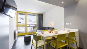 A modern dining area with a white table and yellow chairs, a large window with a view of a snowy landscape, and a flat-screen TV mounted on the wall. - Résidence Le Varet (Bourg-Saint-Maurice)