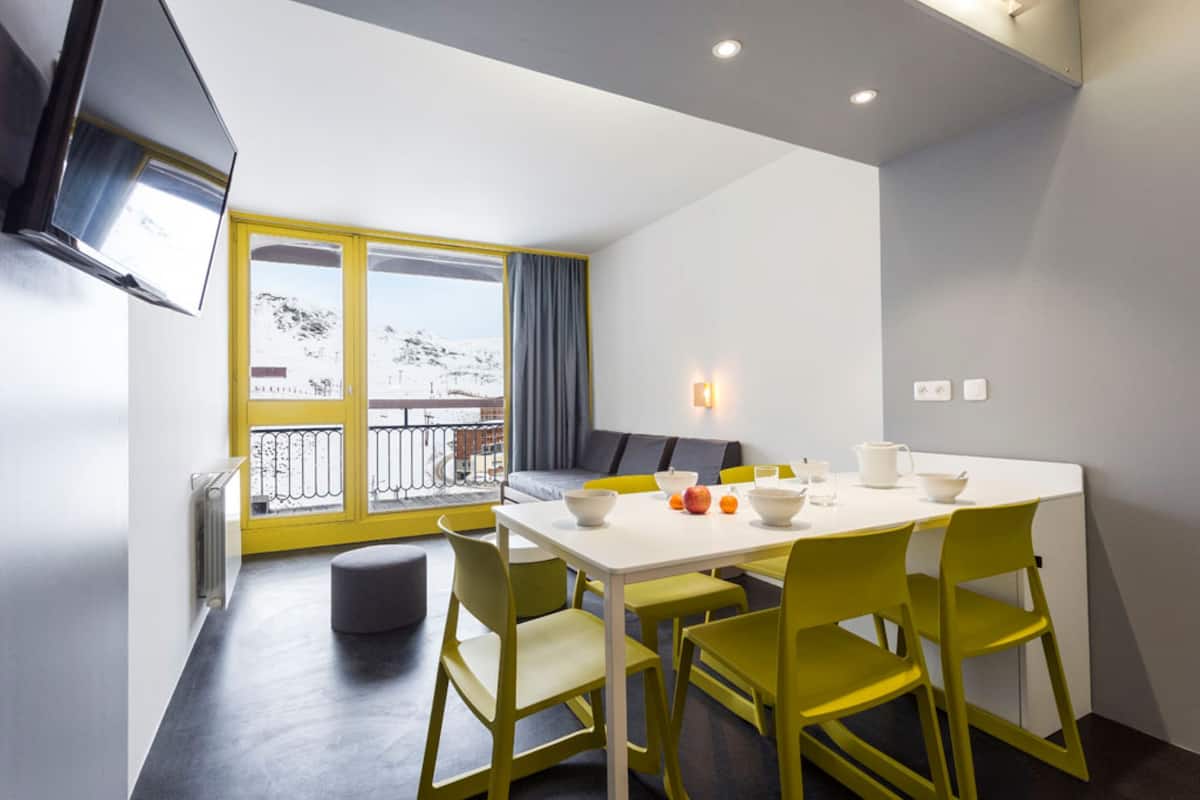 Un espace repas moderne avec une table blanche et des chaises jaunes, une grande fenêtre donnant sur un paysage enneigé et un téléviseur à écran plat fixé au mur.