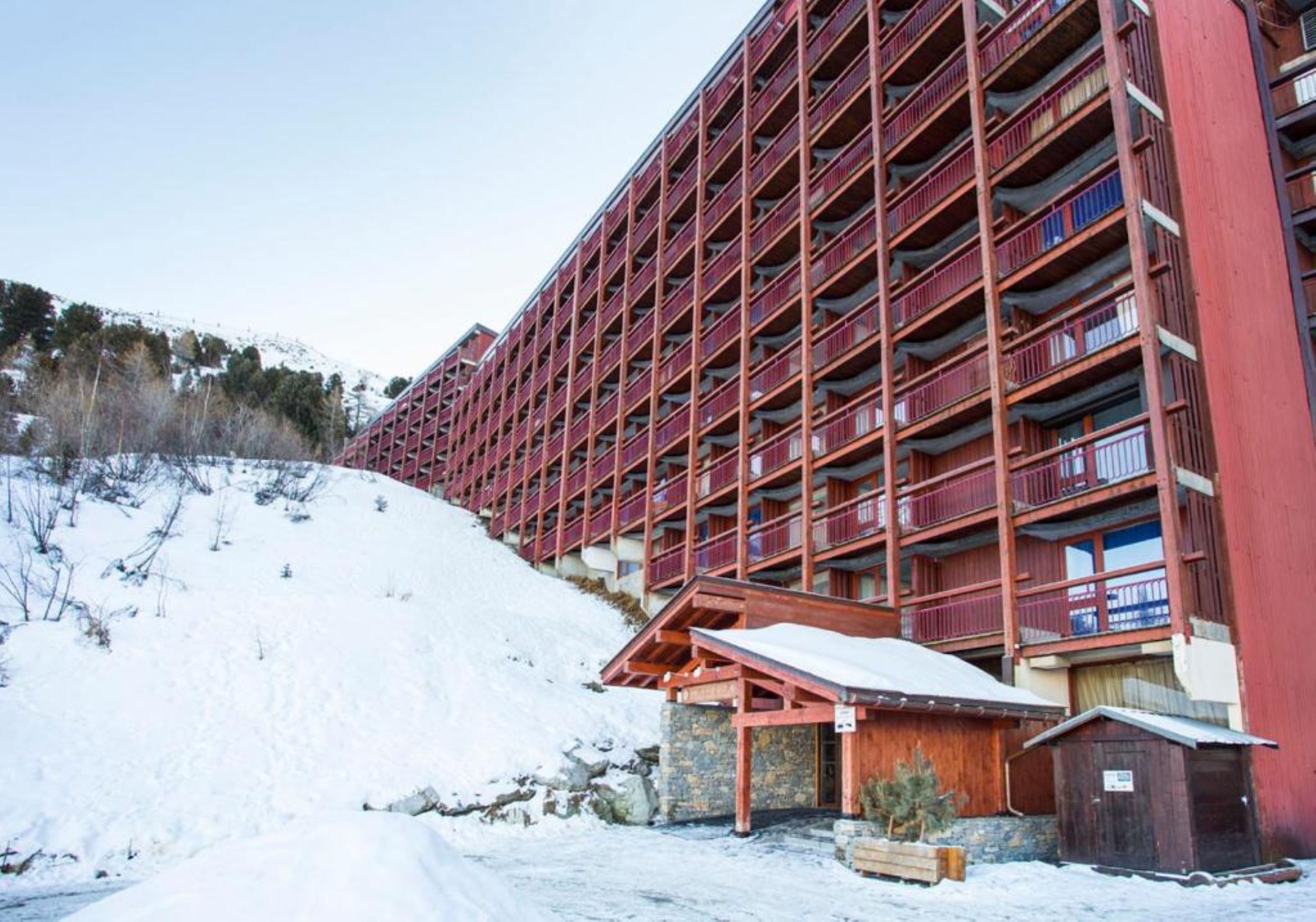 A multi-story hotel with balconies, a ski slope in the background, and a wooden structure at the entrance.
