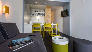 A modern hotel room with a sofa, a small dining table with yellow chairs, a kitchenette, and a flat-screen TV mounted on the wall. - Résidence Le Varet (Bourg-Saint-Maurice)