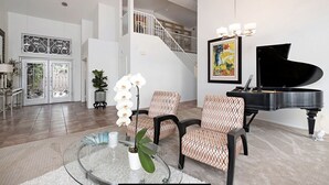 A spacious living room with a grand piano, two patterned armchairs, a glass coffee table with decorative items, and a staircase leading to an upper level. - Large House in Lake La Quinta Community. Gated with Security Guard, Private Pool (La Quinta)