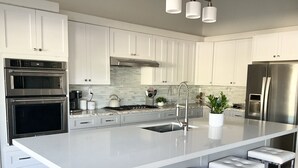A modern kitchen with white cabinets, stainless steel appliances, a marble countertop, and a central island with bar stools. - Large House in Lake La Quinta Community. Gated with Security Guard, Private Pool (La Quinta)