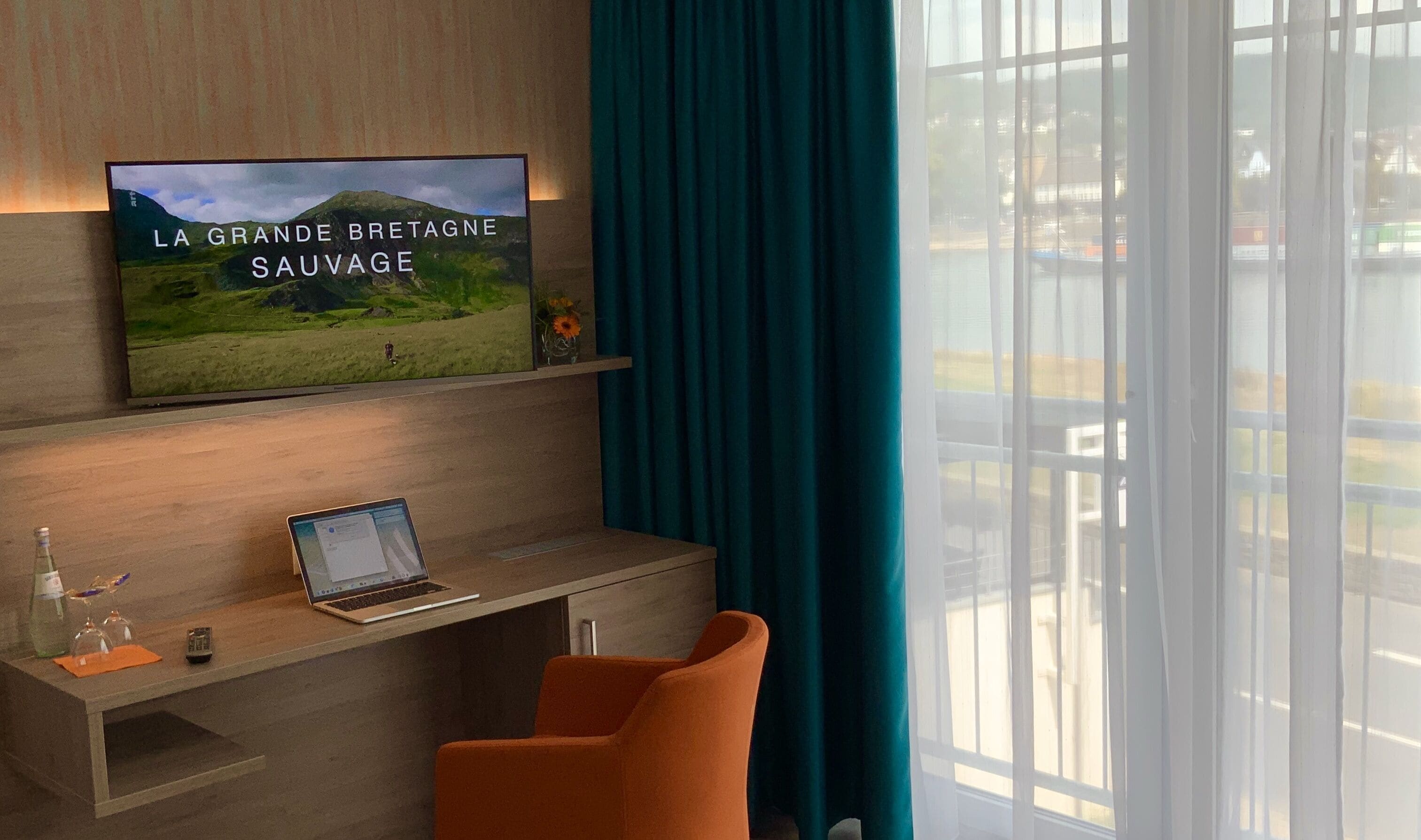 A hotel room with a large window, a laptop on a wooden desk, a TV displaying 'LA GRANDE BRETAGNE SAUVAGE', and a teal curtain.