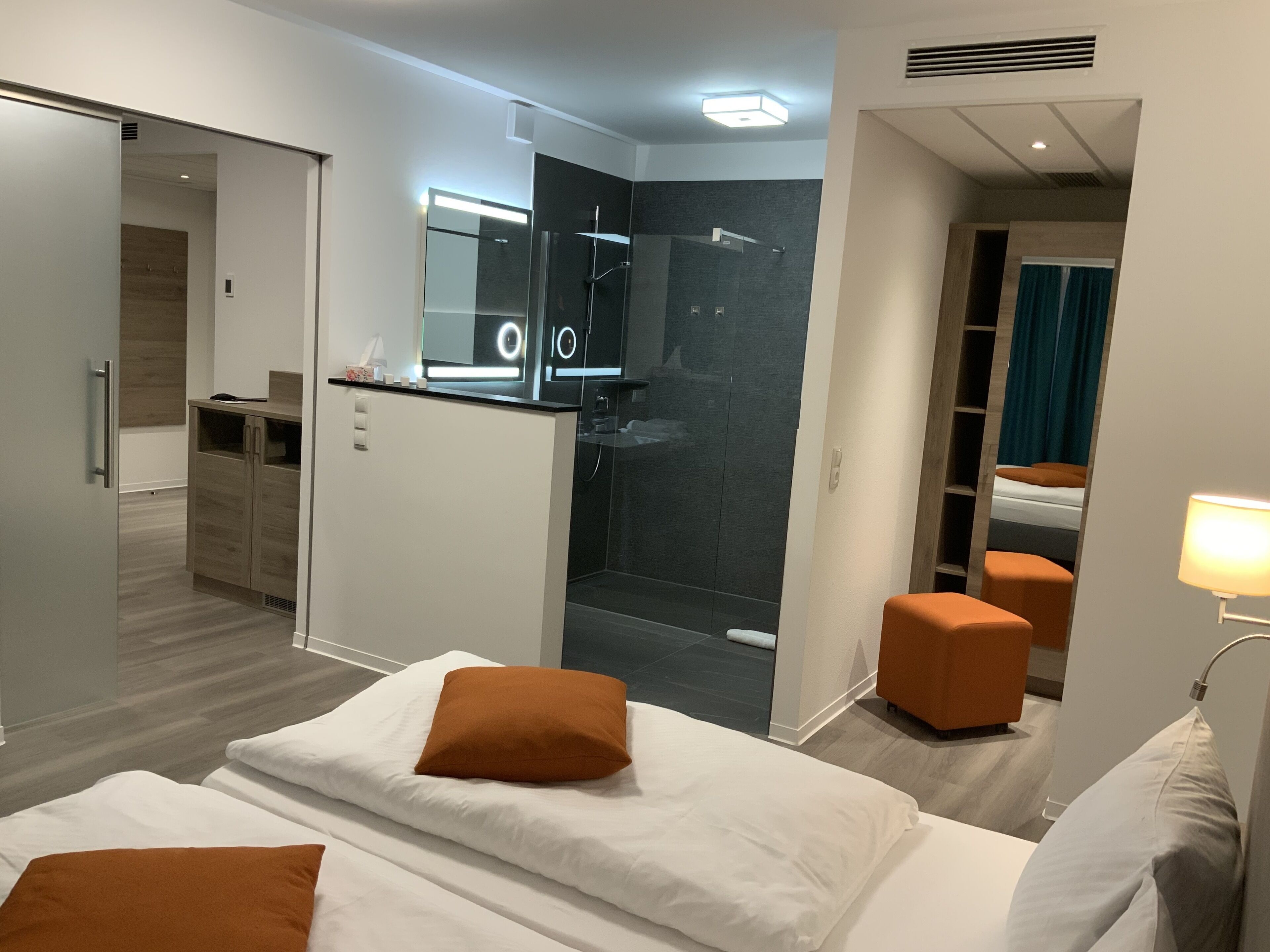 A modern hotel room with a bed, a desk, a shower area, and a sitting area with a lamp.