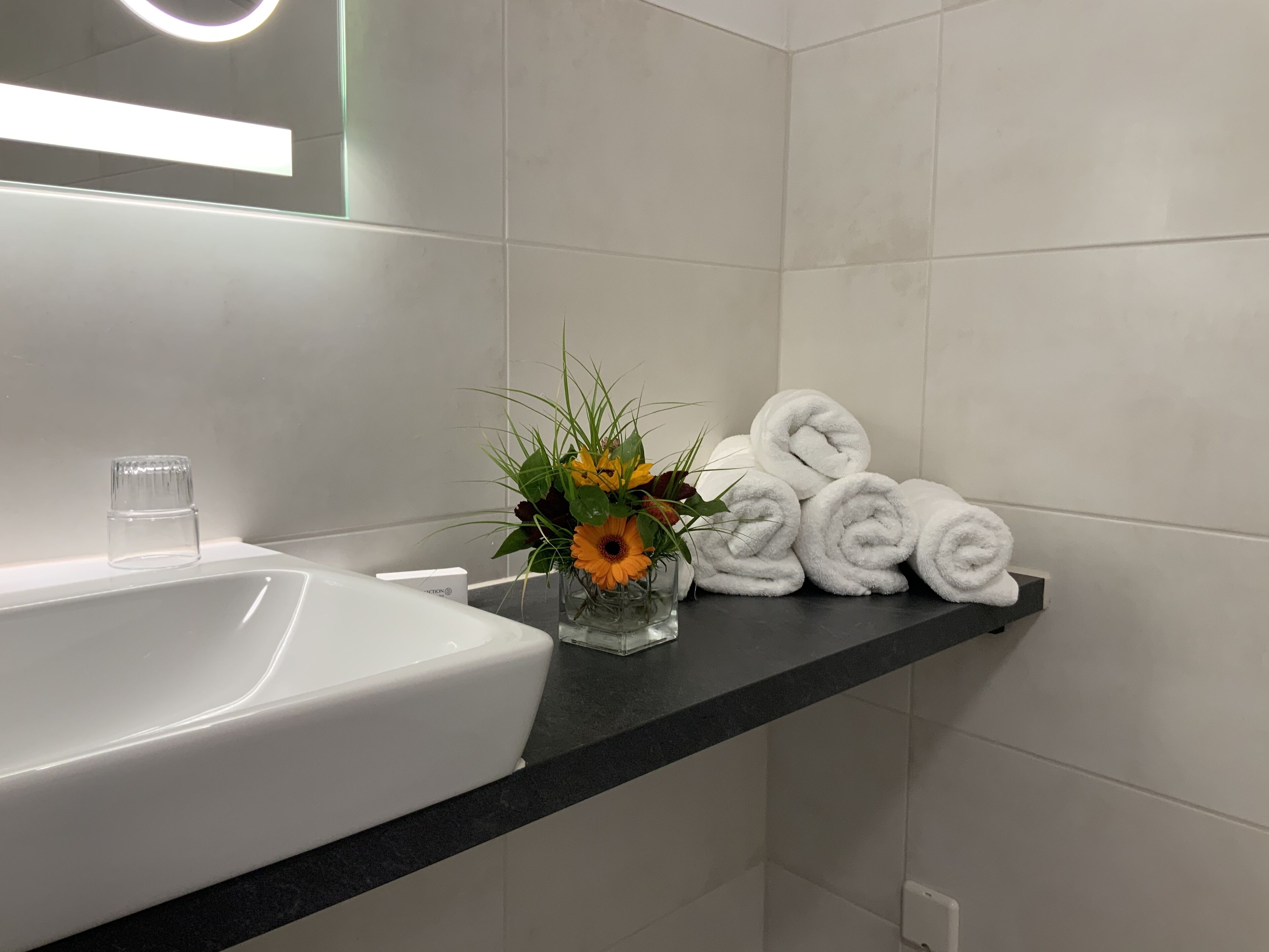 A bathroom with a white sink, a glass vase with flowers, and rolled white towels on a black countertop.