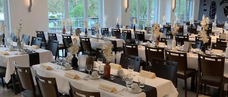 Un comedor amplio con varias mesas dispuestas para un evento formal, con manteles blancos, sillas oscuras y centros de mesa con flores.