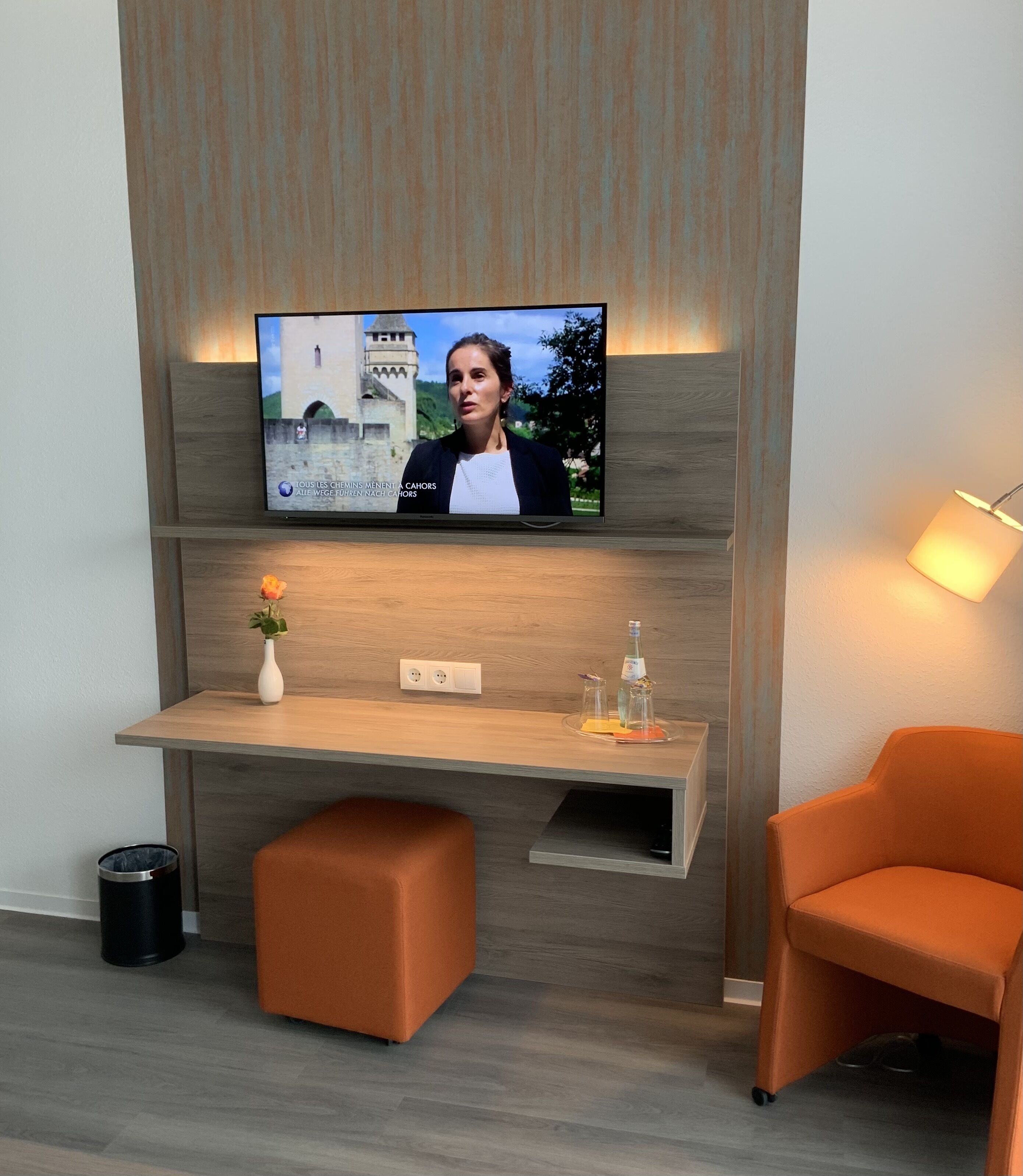 Ein modernes Zimmer mit an der Holzwand montiertem Fernseher, einem kleinen Schreibtisch mit Blumenvase und einem orangefarbenen Sessel.