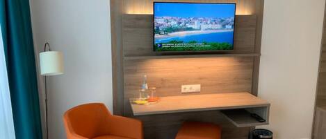 Una sala moderna con un televisor de pantalla plana montado en una pared de madera, una mesita pequeña de madera, dos sillas naranjas y una lámpara de pie.