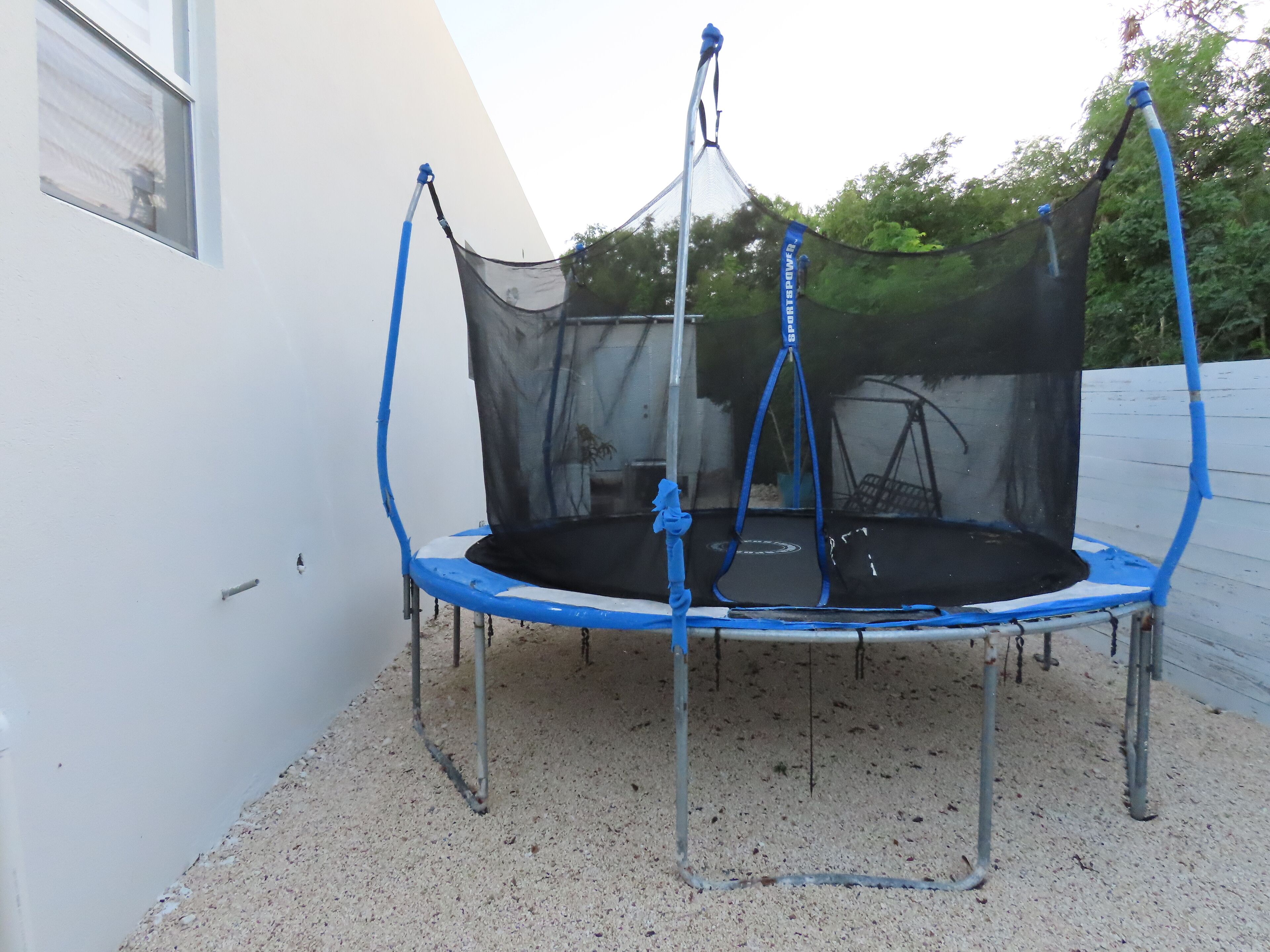 Un trampolín con red de seguridad, sobre una superficie de grava, rodeado por un muro blanco y una cerca de madera.