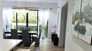 A modern dining area with a long table and chairs, a chandelier, and a large window with curtains. - The White House TCI Guest House (Providenciales)