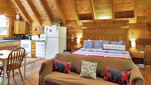 Ein gemütliches Blockhauss-Interieur mit Holzbalkendecke, einer Essecke mit Tisch und Stühlen, einer Küchenzeile und einem Bett mit gemusterten Textilien.