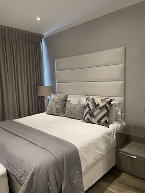 A modern bedroom with a large bed, grey bedding, and a headboard with horizontal lines. - Sandton Smart Apartment Fourteen (Sandton)