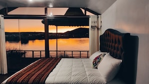 A bedroom with a large bed, a view of a sunset over a body of water, and a balcony with curtains. - Room with Panoramic view in the Uros - Lake Titicaca (Puno)