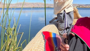 A person standing by a calm lake, holding a long white object, with a woven basket and a colorful fabric nearby. - Room with Panoramic view in the Uros - Lake Titicaca (Puno)