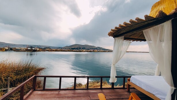 A wooden deck with a bed and a canopy overlooking a calm lake with buildings in the background. - Room with Panoramic view in the Uros - Lake Titicaca (Puno)