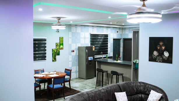 A modern kitchen with a refrigerator, sink, and bar stools. There is a dining area with a table and chairs. - Immaculate 3-bed Apartment in Accra (North Legon)