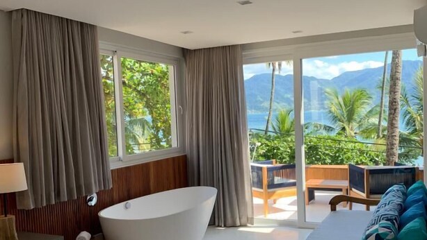 A modern hotel room with a large bathtub, a bed with a wooden frame, and a view of palm trees and mountains through large windows. - Bahari Suites (Ilhabela)