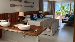 A modern hotel room with a large bed, a wooden vanity with two bowls, a balcony with a view of palm trees, and a wall with decorative paddles. - Bahari Suites (Ilhabela)