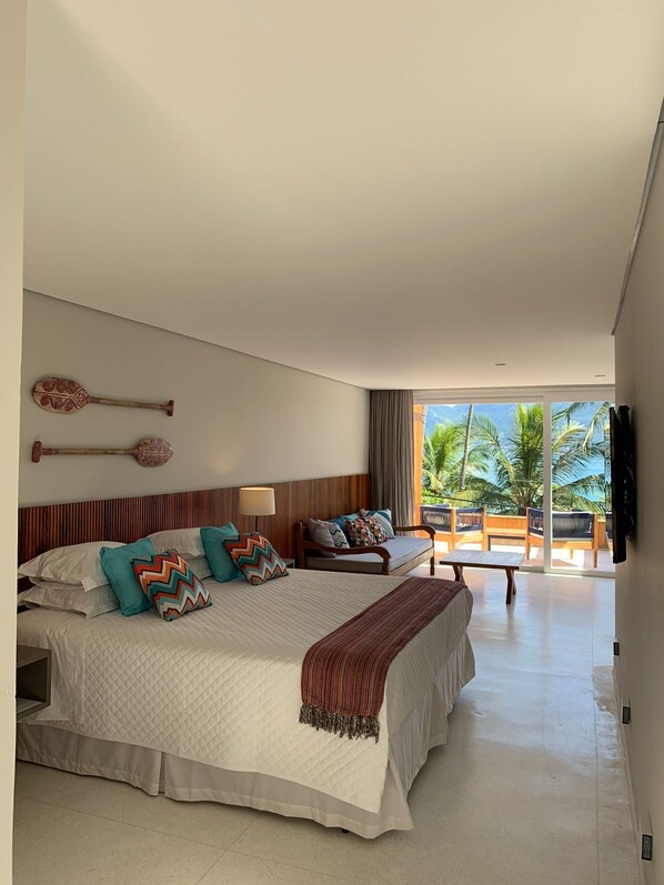 A spacious bedroom with a large bed, a sofa, and a view of palm trees through the window.