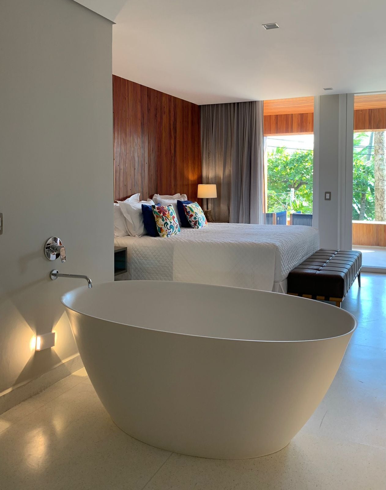 A modern bathroom with a freestanding bathtub, a bed with colorful pillows, and a view of greenery outside.