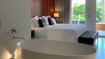 A modern bathroom with a freestanding bathtub, a bed with colorful pillows, and a view of greenery outside.