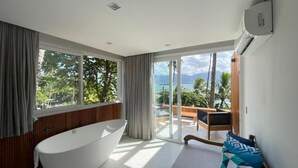 A modern bathroom with a freestanding bathtub, large windows, and a view of a balcony with outdoor furniture. - Bahari Suites (Ilhabela)