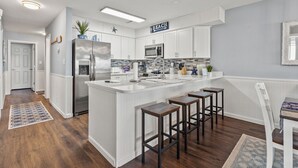 A modern kitchen with white cabinetry, a central island with stools, a sign reading "BEACH", and a microwave above the island. - Updated oceanfront condo w/pool, great location near Main St and other attractions (North Myrtle Beach)