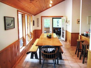 Interior de fusta amb una llarga taula de menjador, seients i cadires. Hi ha finestres i una porta que dóna a l'exterior.