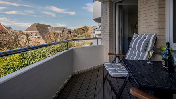 A balcony with a wooden floor, a table with a cushion, a bottle of wine, and a glass.